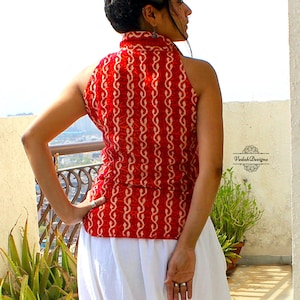 May include: A sleeveless red top featuring a repeating cream-colored vertical vine pattern and a high collar. The garment is paired with a plain white skirt. The text VedahDesigns is visible on the right side of the image near the waistline.