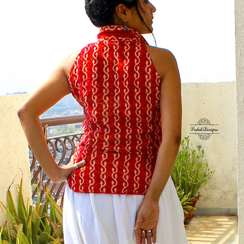 May include: A sleeveless red top featuring a repeating cream-colored vertical vine pattern and a high collar. The garment is paired with a plain white skirt. The text VedahDesigns is visible on the right side of the image near the waistline.