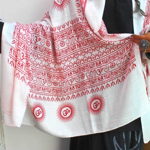 Può includere: Sciarpa bianca con un motivo rosso con simboli del sole e Om. La sciarpa è drappeggiata su un outfit nero. Il design include vari simboli e motivi, creando un look unico e artistico.