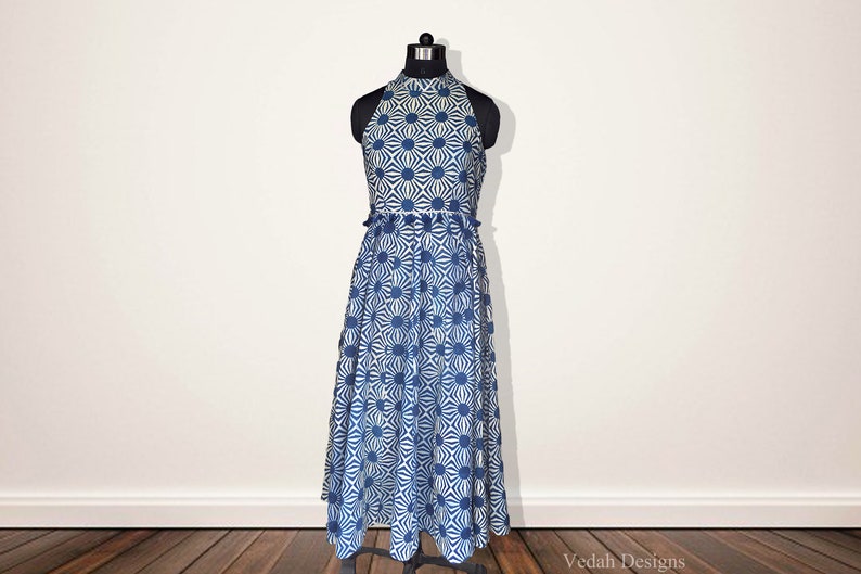 May include: A sleeveless midi dress featuring a high halter neckline and a fitted bodice that transitions into a flared skirt. The fabric is white with a repeating blue geometric sunburst pattern. The text Vedah Designs is visible at the bottom.