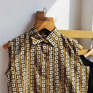 May include: A sleeveless button-down shirt featuring a mustard yellow and white geometric pattern of vertical stripes and squares. The shirt has a pointed collar and a front placket with visible brown buttons, displayed on a wooden hanger.