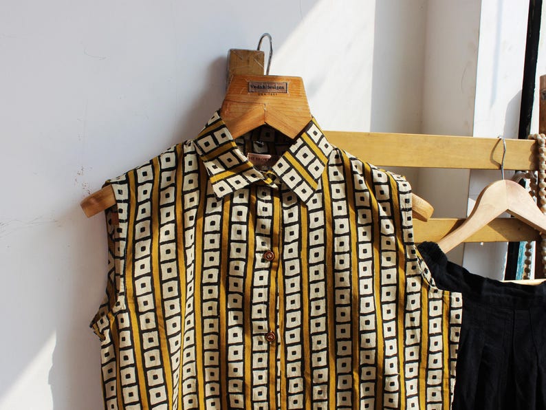 May include: A sleeveless button-down shirt featuring a mustard yellow and white geometric pattern of vertical stripes and squares. The shirt has a pointed collar and a front placket with visible brown buttons, displayed on a wooden hanger.