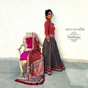 Puede incluir: Una mujer con una blusa a rayas rojas y blancas y una falda larga negra con un estampado de puntos blancos. Un chal estampado en rojo est&aacute; drapeado sobre una silla vintage con un coj&iacute;n rosa. La imagen incluye texto en idioma extranjero.