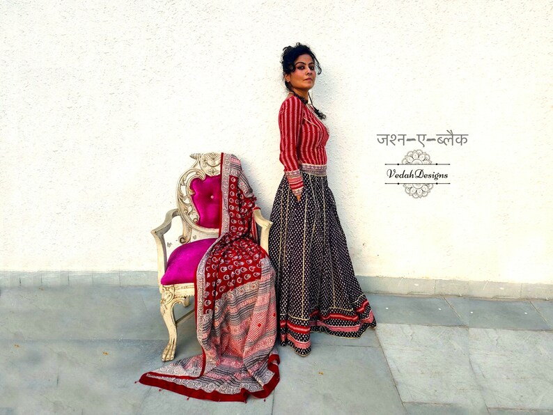 Puede incluir: Una mujer con una blusa a rayas rojas y blancas y una falda larga negra con un estampado de puntos blancos. Un chal estampado en rojo est&aacute; drapeado sobre una silla vintage con un coj&iacute;n rosa. La imagen incluye texto en idioma extranjero.