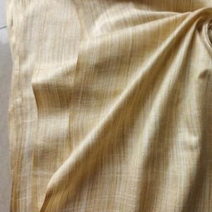 May include: A piece of fabric in a light golden yellow hue featuring a visible vertical slubbed texture. The material is draped to create soft folds and shadows, highlighting the woven grain and the subtle variations in the light yellow color.