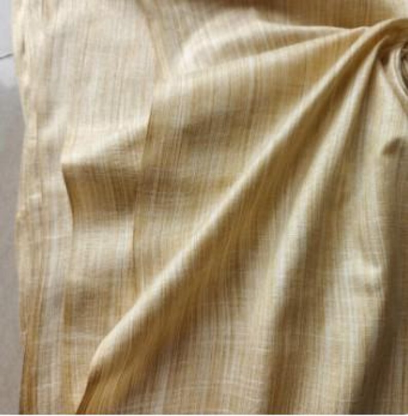 May include: A piece of fabric in a light golden yellow hue featuring a visible vertical slubbed texture. The material is draped to create soft folds and shadows, highlighting the woven grain and the subtle variations in the light yellow color.
