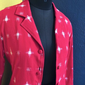 May include: A red blazer featuring a white cross-hatch pattern, a notched lapel, and fabric-covered buttons down the front. The garment is displayed on a black mannequin. The text Vedah Designs is visible in the bottom right corner of the image.
