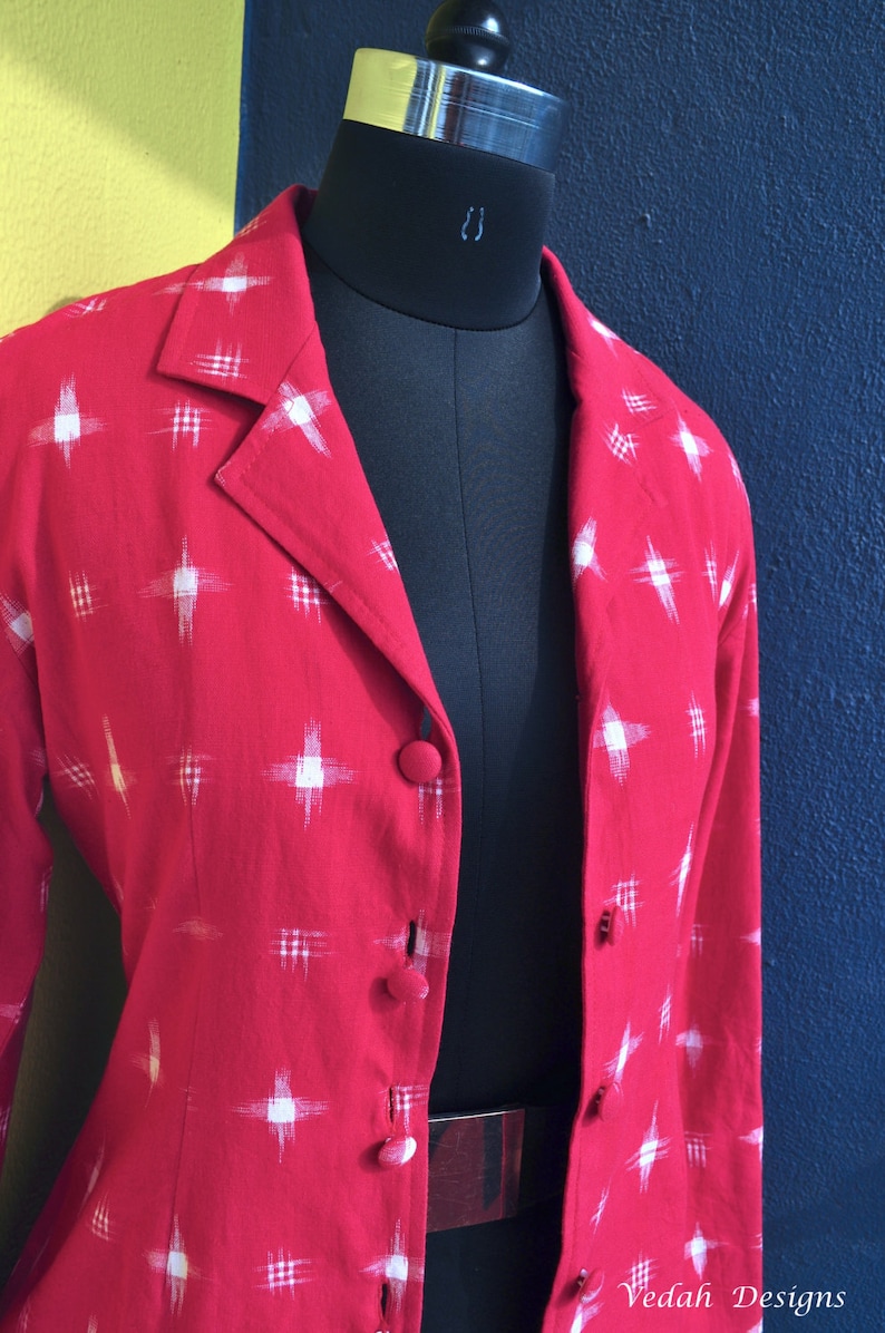 May include: A red blazer featuring a white cross-hatch pattern, a notched lapel, and fabric-covered buttons down the front. The garment is displayed on a black mannequin. The text Vedah Designs is visible in the bottom right corner of the image.