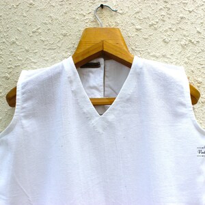 May include: White sleeveless top with a V-neckline hanging on a wooden hanger against a textured white wall.