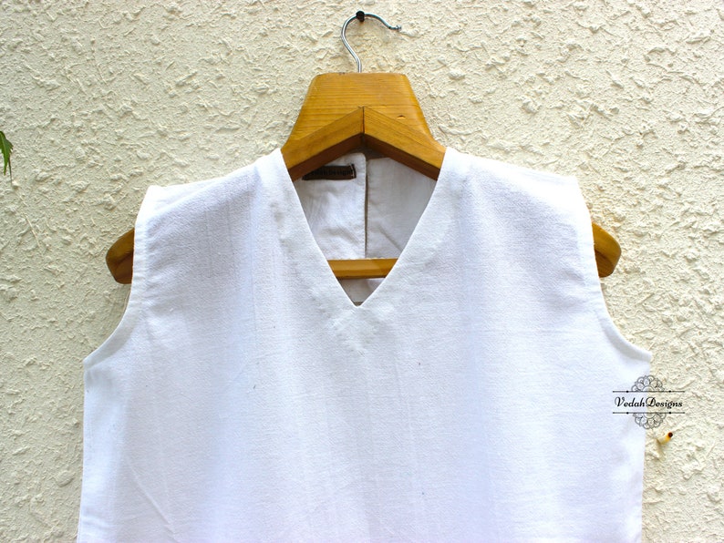 May include: White sleeveless top with a V-neckline hanging on a wooden hanger against a textured white wall.