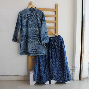 Puede incluir: Un conjunto de dos piezas teñido con índigo, que consta de una túnica de manga larga y una falda fluida. La túnica presenta un cuello mandarín y un patrón geométrico. La falda tiene un patrón a juego y un ajuste amplio y cómodo. El conjunto se exhibe en una percha de madera.