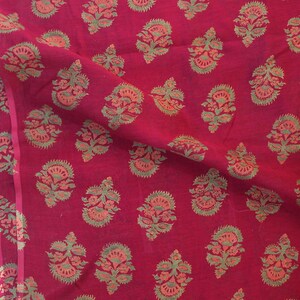 May include: Close-up of a deep red fabric with a repeating floral pattern in shades of green and orange. The fabric is folded, showing the texture and design of the textile. The floral pattern is evenly spaced across the material.