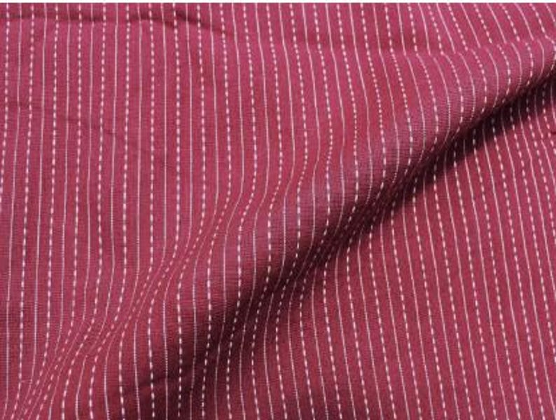 May include: A deep burgundy fabric featuring a pattern of vertical white dashed lines. The material has a visible woven texture and is draped to create soft folds, highlighting the consistent spacing of the white stitch-like markings.