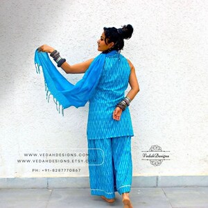 K&ouml;nnte beinhalten: Eine Frau tr&auml;gt ein blaues und wei&szlig;es Ikat-Palazzo-Hosenset mit einem passenden blauen Dupatta. Das Outfit besteht aus einem &auml;rmellosen Oberteil mit hohem Ausschnitt und einer passenden Palazzo-Hose mit weitem Bein. Der Dupatta ist ein hellblauer, durchsichtiger Stoff mit Fransenbesatz. Die Frau steht vor einer wei&szlig;en Wand mit dem Text "www.vedahdesigns.com" und "www.vedahdesigns.etsy.com", der auf die Wand gedruckt ist. Der Text "Vedah Designs" ist ebenfalls in einem Kreis auf die Wand gedruckt.