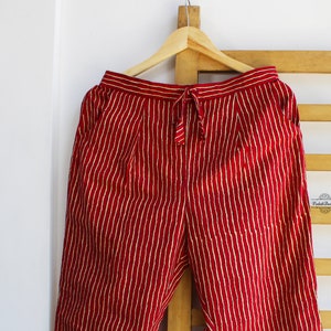 May include: Red trousers featuring a vertical white pinstripe pattern, an elasticized waistband with a matching drawstring tie, and side pockets. The garment is displayed on a wooden hanger, showcasing a straight-leg cut and a classic design.