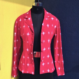 May include: A long-sleeved red blazer featuring a white cross-hatch and star-like pattern. The blazer has a notched lapel collar, a front button closure with matching red buttons, and is displayed on a black mannequin torso.