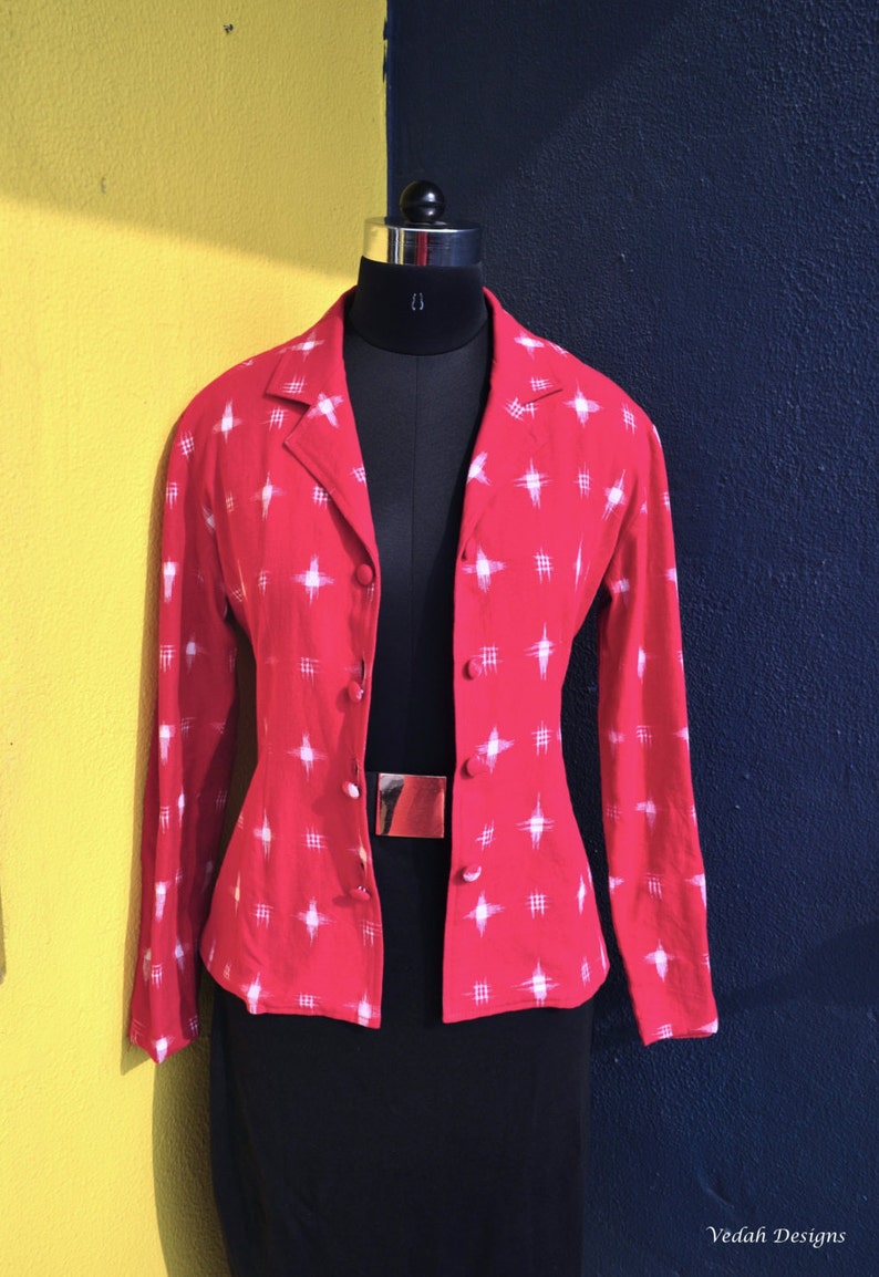 May include: A long-sleeved red blazer featuring a white cross-hatch and star-like pattern. The blazer has a notched lapel collar, a front button closure with matching red buttons, and is displayed on a black mannequin torso.