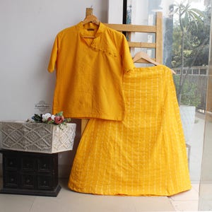May include: A bright yellow two-piece set consisting of a short-sleeved top with a mandarin collar and side button closure, paired with a matching long skirt featuring vertical white stripes. The set is displayed on wooden hangers.