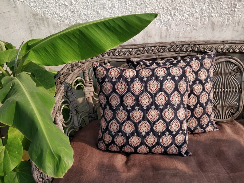 May include: Two black and white patterned throw pillows with red and cream accents are placed on a brown wicker chair. A large green leaf from a plant is in the foreground.