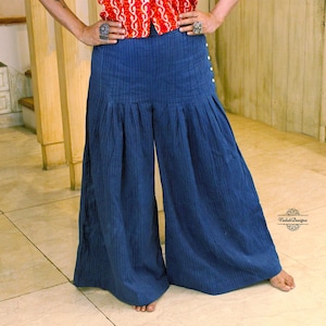 May include: High-waisted wide-leg trousers in a dark blue pinstripe fabric. The pants feature a series of small white buttons along the left side hip and a pleated design that creates a voluminous silhouette. The fabric has a subtle vertical texture.
