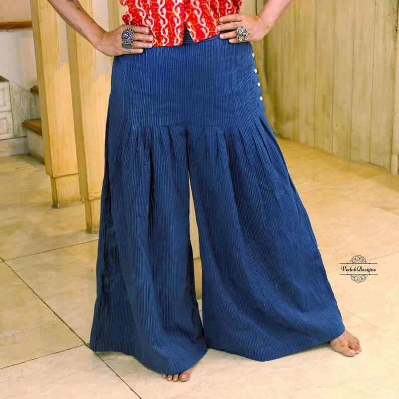 May include: High-waisted wide-leg trousers in a dark blue pinstripe fabric. The pants feature a series of small white buttons along the left side hip and a pleated design that creates a voluminous silhouette. The fabric has a subtle vertical texture.