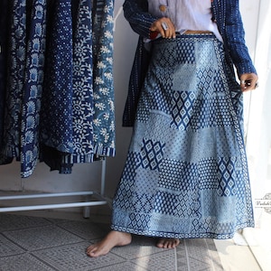Puede incluir: Una falda larga de corte en A con un diseño de patchwork de varios patrones geométricos en azul índigo y blanco. La falda tiene una cintura alta y una silueta fluida. Un pequeño logotipo con el texto Vedah Designs es visible en la esquina inferior derecha.