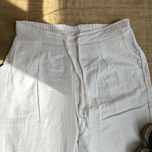 May include: White pants featuring an elasticized waistband with a drawstring closure. The garment includes two side pockets and a textured fabric finish. The design includes vertical pleats extending from the waistline down the front of the legs.