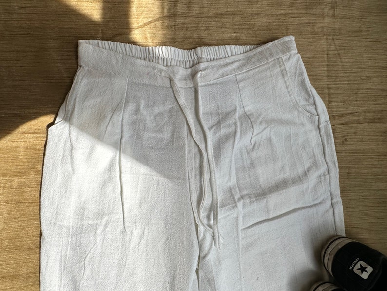May include: White pants featuring an elasticized waistband with a drawstring closure. The garment includes two side pockets and a textured fabric finish. The design includes vertical pleats extending from the waistline down the front of the legs.