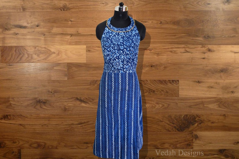 May include: A blue halter dress with a floral and striped pattern. The top section features a floral design, while the lower part has vertical stripes. The dress is displayed on a black mannequin against a wooden background. The dress has a gold beaded neckline.