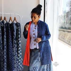 Puede incluir: Una chaqueta azul índigo de manga larga con un patrón de puntos blancos, cuello mao y forro interior naranja. La chaqueta se lleva sobre una camisa azul claro con botones de madera grandes y una falda azul estampada.