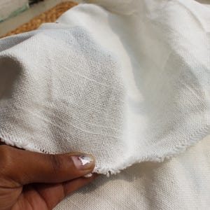 May include: A piece of off-white fabric with a visible woven texture and frayed, raw edges. The material appears lightweight and semi-sheer, showing a loose weave pattern throughout the surface of the textile, which is folded to display its drape.