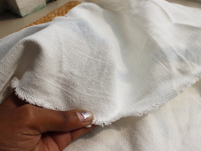 May include: A piece of off-white fabric with a visible woven texture and frayed, raw edges. The material appears lightweight and semi-sheer, showing a loose weave pattern throughout the surface of the textile, which is folded to display its drape.
