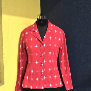 May include: A long-sleeved red blazer featuring a white cross-hatch pattern throughout. The garment has a notched lapel collar and a front button closure with four matching red fabric-covered buttons. The blazer is displayed on a mannequin.