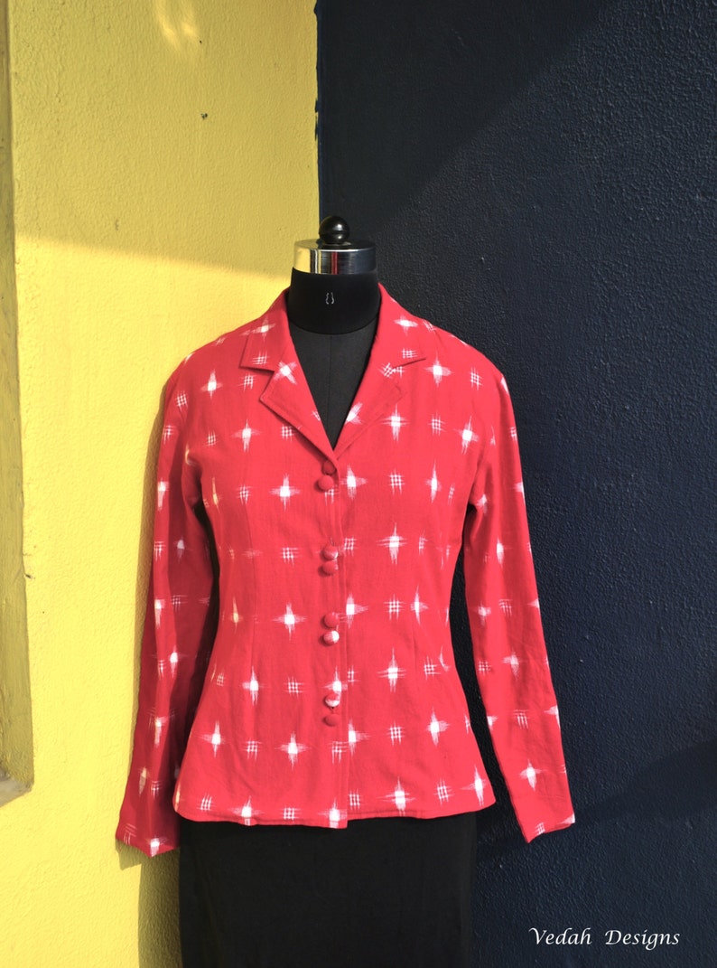 May include: A long-sleeved red blazer featuring a white cross-hatch pattern throughout. The garment has a notched lapel collar and a front button closure with four matching red fabric-covered buttons. The blazer is displayed on a mannequin.