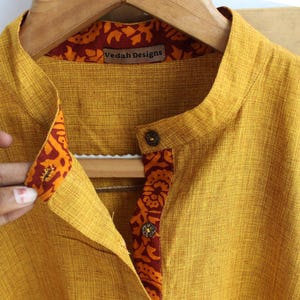 May include: Mustard yellow shirt with a mandarin collar and decorative patterned trim. The shirt has a button closure and the label "Veda Designs" is visible. The trim features a floral design in shades of orange and red.