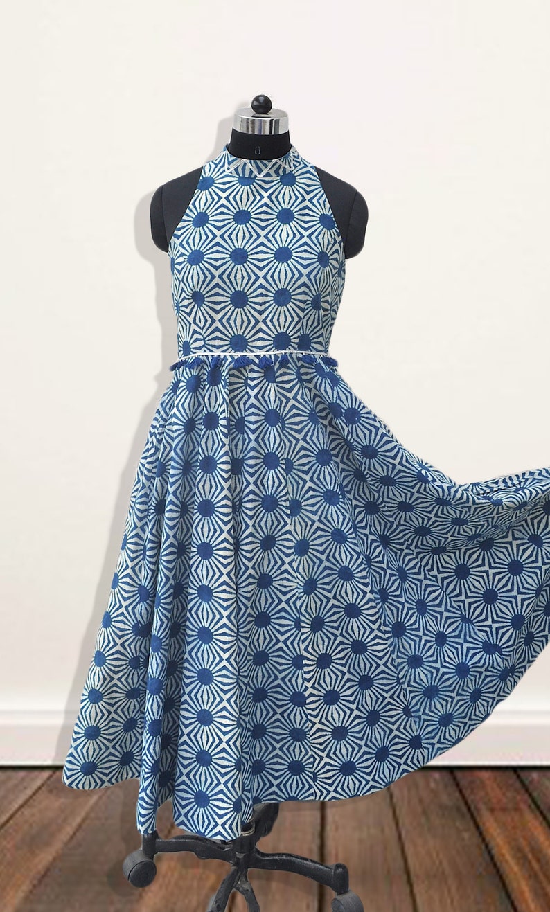 May include: A sleeveless midi dress featuring a high neckline and a flared skirt. The garment is decorated with a repeating blue and white geometric sunburst pattern. A thin waistband with small blue tassels separates the bodice from the skirt.