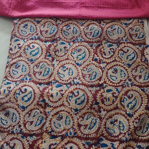 Op de afbeelding: Een textielstof met een herhalend cirkelvormig paisley-patroon in tinten dieprood, blauw en cr&egrave;me. De stof ligt onder een strook effen magentakleurige stof met dunne, horizontale witte krijtstrepen langs de bovenrand.
