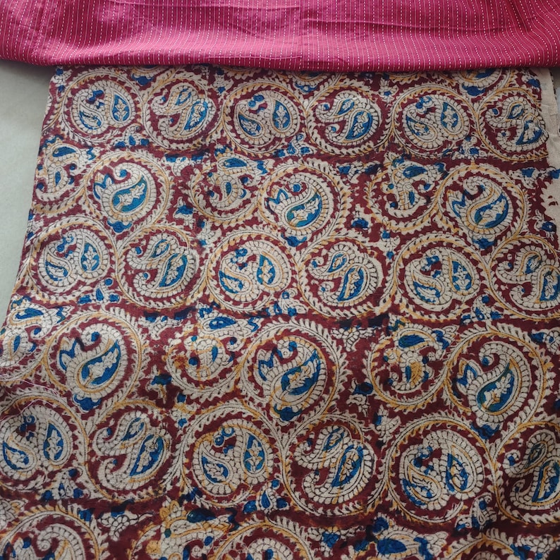 Op de afbeelding: Een textielstof met een herhalend cirkelvormig paisley-patroon in tinten dieprood, blauw en cr&egrave;me. De stof ligt onder een strook effen magentakleurige stof met dunne, horizontale witte krijtstrepen langs de bovenrand.