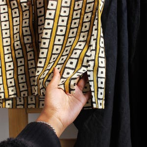 May include: A button-down shirt featuring a mustard yellow and white vertical striped pattern with black square outlines, paired with a solid black skirt. The shirt has a small side slit at the hem and brown buttons along the front placket.