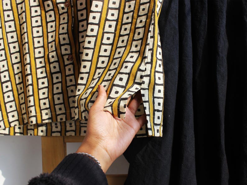 May include: A button-down shirt featuring a mustard yellow and white vertical striped pattern with black square outlines, paired with a solid black skirt. The shirt has a small side slit at the hem and brown buttons along the front placket.