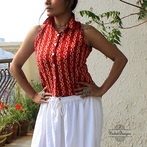 May include: A sleeveless red collared shirt featuring a white vertical wavy pattern and a front button closure, paired with a white skirt that has an elastic waistband with a drawstring. The text VedahDesigns is visible in the bottom right corner.