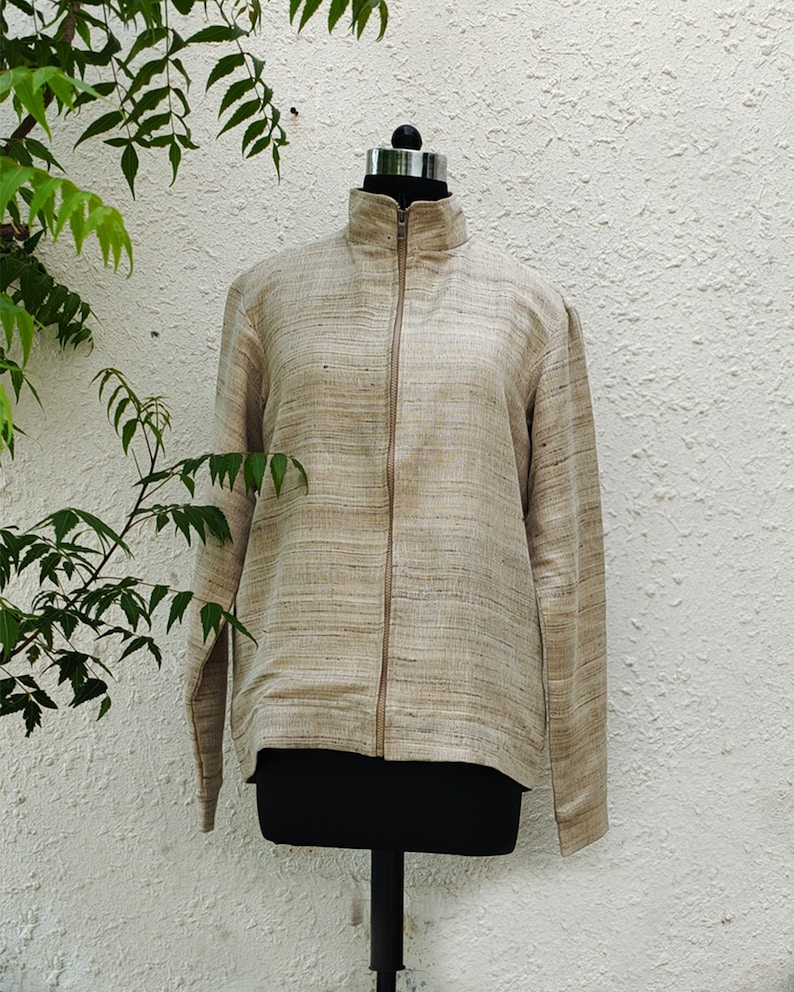 May include: A long-sleeved jacket in a light tan and beige textured fabric with a visible horizontal weave pattern. The jacket features a high collar, a full-length front zipper closure, and straight sleeves, displayed on a black mannequin torso.