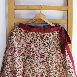 Op de afbeelding: Een wikkelrok met een cr&egrave;mekleurige basis en een dicht bloemenpatroon in rood- en zwarttinten. De rok heeft een brede, getextureerde bordeauxrode tailleband met een bijpassende striksluiting aan de zijkant, hangend aan een houten kledinghanger.