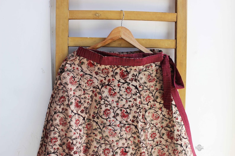 Op de afbeelding: Een wikkelrok met een cr&egrave;mekleurige basis en een dicht bloemenpatroon in rood- en zwarttinten. De rok heeft een brede, getextureerde bordeauxrode tailleband met een bijpassende striksluiting aan de zijkant, hangend aan een houten kledinghanger.