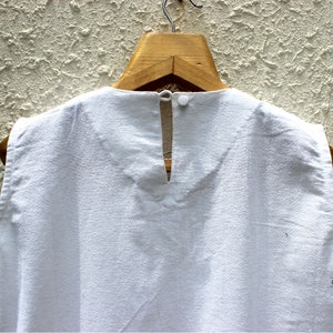 May include: White sleeveless top with a keyhole back opening. The top is hanging on a wooden hanger against a textured wall.