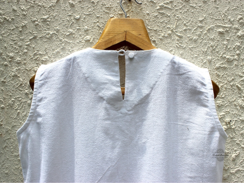 May include: White sleeveless top with a keyhole back opening. The top is hanging on a wooden hanger against a textured wall.