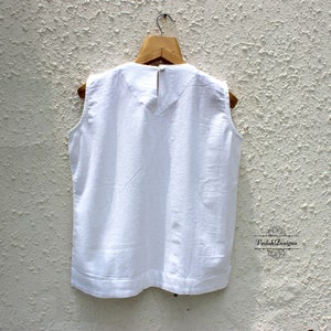 May include: White sleeveless top with a round neckline. The top is hanging on a wooden hanger against a white textured wall.