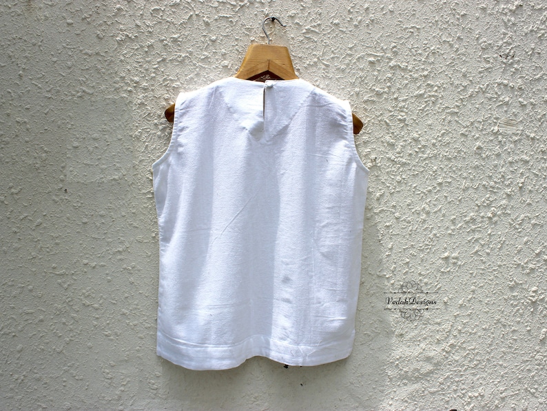 May include: White sleeveless top with a round neckline. The top is hanging on a wooden hanger against a white textured wall.