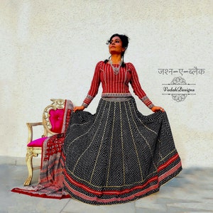 Puede incluir: Una mujer con un atuendo tradicional indio. El conjunto incluye una parte superior de manga larga a rayas rojas y blancas y una falda negra con patrones de puntos y rayas blancas. El texto "Jashn-e-Black" y "Veda Designs" son visibles.
