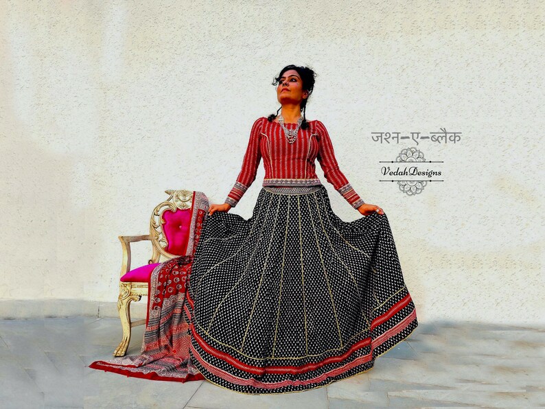 Puede incluir: Una mujer con un atuendo tradicional indio. El conjunto incluye una parte superior de manga larga a rayas rojas y blancas y una falda negra con patrones de puntos y rayas blancas. El texto "Jashn-e-Black" y "Veda Designs" son visibles.